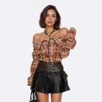 A model wearing a brown and beige plaid off-the-shoulder zip-up jacket with dramatic puffed sleeves, paired with a black faux-leather mini skirt and a black beaded necklace, against a plain white background.