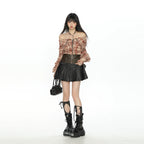 A model stands against a solid white studio background wearing a plaid off-the-shoulder crop top, a black faux-leather pleated mini skirt, a brown leather waist belt, black lace-up combat boots, and holding a small black handbag.