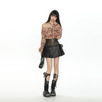 A model stands against a solid white studio background wearing a punk/goth style outfit consisting of a brown and red plaid off-the-shoulder long-sleeve top, a black faux-leather corset, a black pleated mini skirt, and black lace-up combat boots.