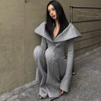 A woman with long black hair sits on a concrete ledge against a textured beige wall, wearing a light gray knit matching set consisting of a wide collar coat and wide-leg trousers.