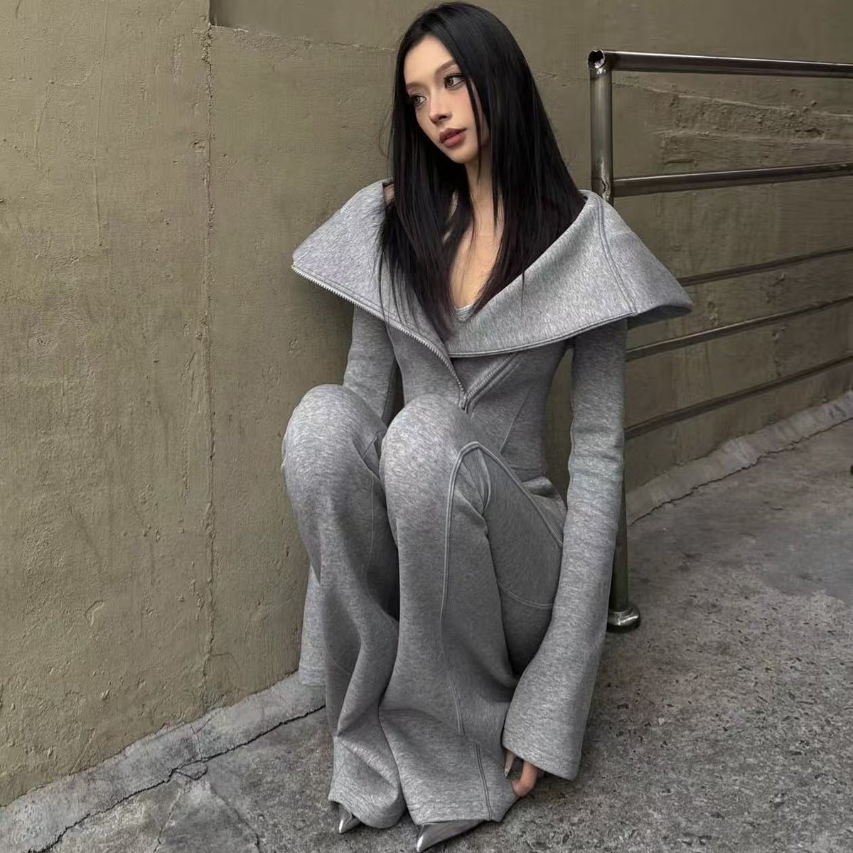 A woman with long black hair sits on a concrete ledge against a textured beige wall, wearing a light gray knit matching set consisting of a wide collar coat and wide-leg trousers.