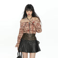 A model wearing a brown and beige plaid off-the-shoulder long-sleeve top with a front zipper, paired with a black faux-leather pleated mini skirt and a dark brown corset-style waist panel.