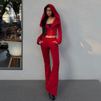 A woman wearing a bright red two-piece outfit consisting of a cropped zip-up hoodie with a matching hood and matching flared pants, standing against a light gray wall.