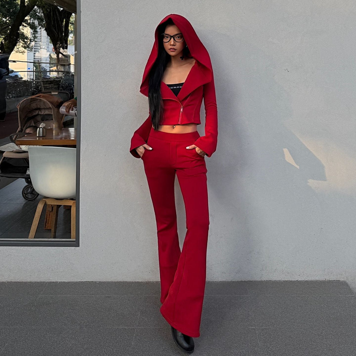 A woman wearing a bright red two-piece outfit consisting of a cropped zip-up hoodie with a matching hood and matching flared pants, standing against a light gray wall.