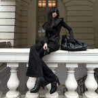A model wearing a black long-sleeve top, black wide-leg pants, and black boots sits on a white stone balustrade. A large black crocodile-embossed handbag rests beside her on the ledge.